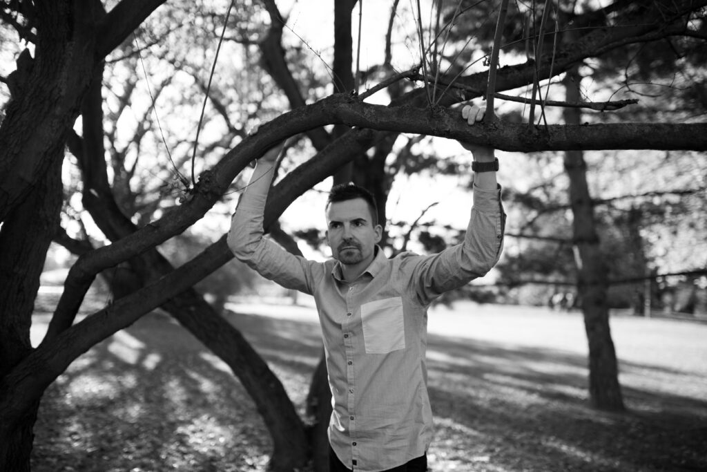 Peter Schröter steht in einem Park unter einem knorrigen Baum, beide Hände an einem Ast über dem Kopf, und blickt zur Seite.