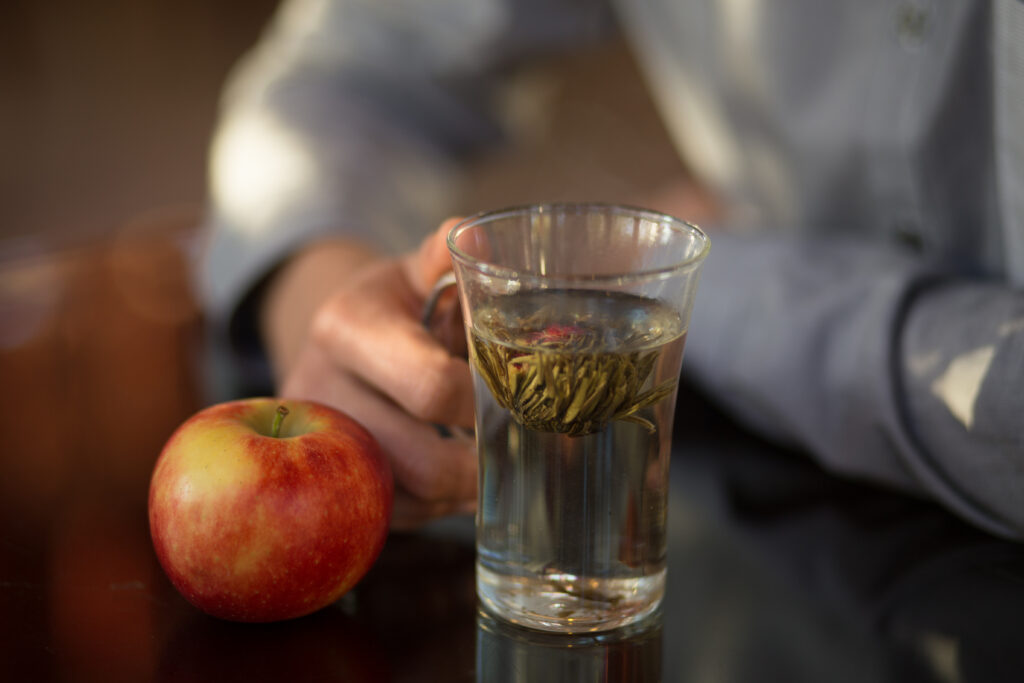 Ein Glas Tee in der Hand, daneben ein roter Apfel auf einem dunklen Tisch.