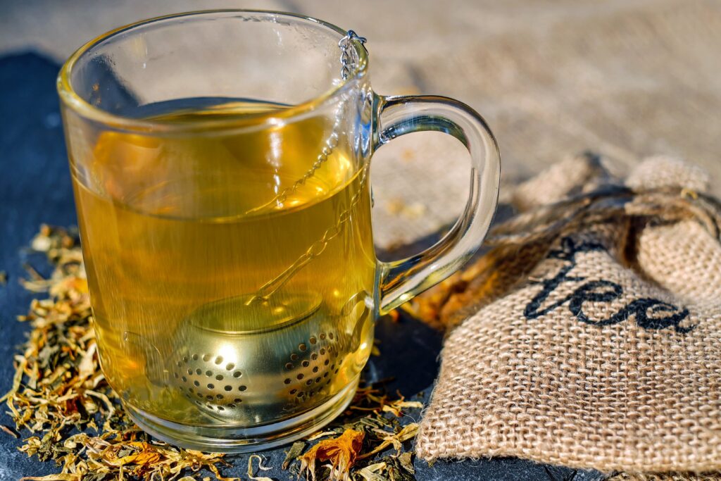 Glas mit goldgelbem Kräutertee und Teesieb, daneben ein Jutesack mit der Aufschrift Tee und getrocknete Kräuter.